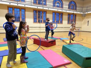 Five young people in masks play with hoops and mats in a gym.