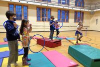 Five young people in masks play with hoops and mats in a gym.