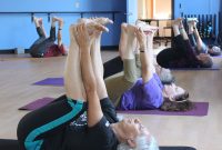 Six women lay on their backs on mats in a studio practicing a leg stretch.